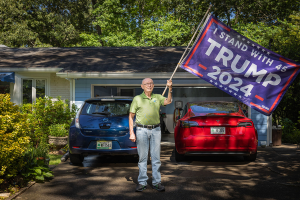 Don Francis waves a “Trump 2024” flag in the driveway of his home next to two electric vehicles.